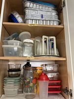 Wide view of wooden cabinet shelves, showing aluminum cooking pans on top shelf, assorted plastic containers stacked, vintage floral glass jar, plastic shaker with red lid, yellow Thermoflask and other storage items.