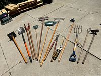 Photo showing full spread of various gardening tools including rakes, shovels, hoes, a pitchfork, and loppers laid out on pavement