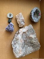 Top down view showing six geode and mineral specimens arranged in a cardboard box, highlighting the large clear crystal cluster and partially hollow geodes.