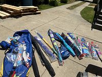 Multiple kites and two large blue carrying cases laid out on pavement, showing variety of kite sizes and packaging.