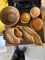 Top-down photo showing six wooden serving pieces including bowls and trays arranged on a dark surface, showcasing their natural wood grain and mid-century modern design.