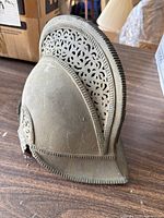 Side view showing the dusty gray metal helmet with ornate floral cut-out patterns along the crest and embossed decorative edges.