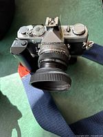 Front view showing Olympus OM-1 branding, lens with hood attached, and battery grip attached to the base. Camera strap in navy blue color visible.