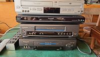 Stack of four Panasonic devices including a silver DVD player, a black Blu-ray player, and two black VCRs shown from the front.