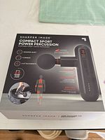 Box front showing product name 'Sharper Image Compact Sport Power Percussion' and key features such as whisper quiet, 4 speeds, 4 attachments, high intensity, rechargeable with 2 hour runtime, compact design.