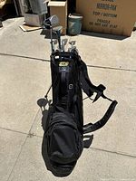Front view of black stand golf bag containing clubs, standing upright on concrete.
