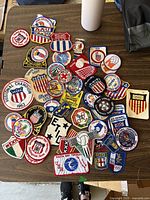 Over 40 varied sports patches from 1960s-70s laid out on a table, showing range of colorful embroidery and sports themes including volleyball, soccer, and Olympic patches.