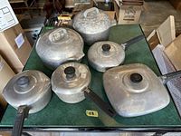 Top view showing full set of 7 Wagner Ware Magnalite pots and pans with lids on a green surface