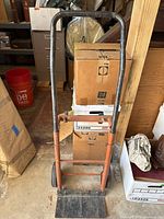 Front view of orange metal hand truck showing black handles, loading base and metal wheels, background includes boxes and household items.