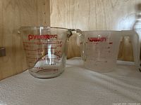 Two glass measuring cups, one Pyrex branded, with red measurement markings and handles on wooden shelf.