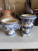 Two vintage Dutch ceramic vases featuring traditional blue and white floral patterns, placed on a shelf among other objects.