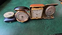 Front view of four vintage travel alarm clocks and one pocket watch arranged side by side on green surface, showing fold-up cases and different designs.