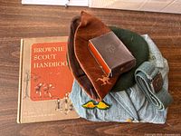 Photo showing vintage Brownie Scout uniform top, patches, Belt, Brownie hat, Brownie Scout Handbook, and a small boxed item.