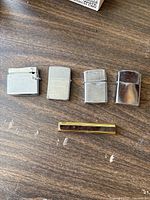 Five vintage lighters arranged on a wooden surface: four silver-tone metal lighters and one slim gold-tone lighter.