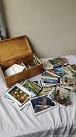 Wooden box filled with assorted vintage postcards spread out on a cloth surface, showing images of landscapes, buildings, and cultural scenes.