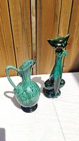 Two green glazed ceramic pieces on a white surface, cat figurine and pitcher vase shown upright against wood background.