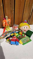 Full view of the collection showing the orange transparent robot, character face toy, green box, blue-red toy truck, maze game, balls, toy guns, and miscellaneous items on white background with wooden wall.