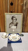 Framed black and white photo of Princess Diana, commemorative plate on blue stand, and matching teacup and saucer with Royal Family crests arranged on white cloth in front of wooden backdrop.