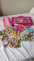 Photo showing bright pink Shopkins storage case, numerous small bags filled with multicolored miniature Shopkins toys, pink miniature shopping cart, and other small toy accessories arranged on white fabric.