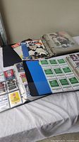 Photo showing three binders laid out, two with trading cards visible inside clear protective sheets, and one mostly closed. Card types include hockey and basketball trading cards, some loose cards, and unopened card packs also present.