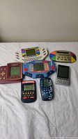 Group of six vintage handheld electronic games laid out on white cloth, showing different shapes, colors, screen types, and button layouts.