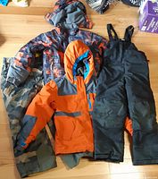 Four outerwear pieces laid out: two jackets, camo rain pants, black bib snow pants