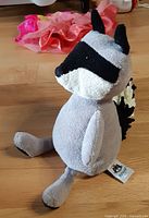 Side view of grey Jellycat raccoon plush seated on wood floor