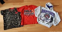 All three Toronto Raptors tops laid out: camo tee, red tee, grey hoodie