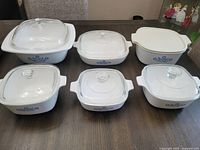 Set of six Corning Ware casserole dishes with Blue Cornflower design and glass lids arranged on table.