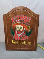 Closed front view of wooden dartboard cabinet with colorful Kings Head Pub & Lodging painting on doors