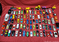Wide shot of 63 die cast cars including Hot Wheels and other brands arranged on red cloth
