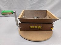 Side angle of the JSP malt mill showing wooden hopper, yellow adjustment knob, and green-handled crank