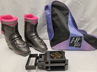 Pair of black Nordica Bioflex women's ski boots with pink interior lining, purple and black ski boot bag, and black plastic accessories on a white backdrop