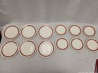 Top view of two rows of plates - 8 large dinner plates and 6 smaller plates with red, gold, and leaf design around rim on white background.