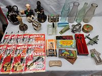 Wide view of entire table showing diverse vintage collectibles: glass bottles, figurines, polishing cloths, small toys, games, and tools arranged on a white table cover.