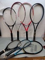 Eight used tennis rackets of various colors and brands leaning against a white background, visible wear on grips and strings.