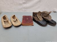 Photo showing two pairs of vintage baby shoes and a small leather-bound book on a gray surface.