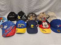 Ten vintage baseball caps arranged in two rows on a gray background showing a variety of colors, logos, and embroidery styles including racing, cartoon characters, and brand logos.