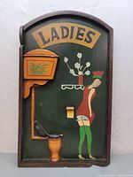 Full view of vintage ladies restroom sign, showing painted figure, toilet, and flower shelf detail.