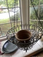 Hanging wrought iron planter with decorative scalloped rim containing a round rustic metal bowl, alongside a long black wooden dough bowl and a black metal bowl on the window sill.