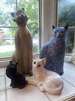 Seven ceramic cat figurines displayed on a window ledge with natural lighting showing different colors and sizes, including beige tall cat, blue speckled cat, white lying cat, and several black cats.