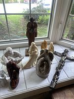 Nine assorted religious statues placed on a windowsill with outdoor light, showing different styles and colors