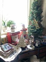 Photo showing lot items on dresser near window: small green artificial Christmas tree with decorations, vintage white ceramic Christmas tree, snow globe, Santa Claus themed box, dishes and cutlery.