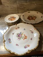 Photo showing one large plate with floral patterns and gold scalloped edges, one medium plate with fruit and floral designs, and four smaller plates stacked with similar decorative style.