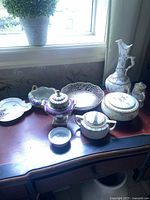 Wide view of two vintage porcelain items on a wooden table near a window: a lidded dish and a pitcher with floral designs and gold detailing.