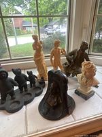 Photo showing assorted vintage small statues on a windowsill, featuring metal Shakespeare bookend, metal man on bench, resin nude statues, classical bust, and set of four small figurines.