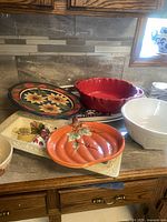 Overview of all dishes together on countertop: colorful floral tray, red serving dish, white footed bowl, pumpkin-shaped dish, rectangular vegetable platter; shows variety and condition