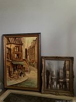 Pair of 2 original framed paintings with urban street scenes, one larger and one smaller, both in frames, positioned against a wall