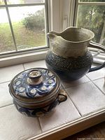 Photo showing the pair of pottery pieces on a white tiled surface near a window, including the large jug and the lidded dish.