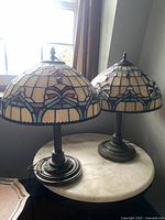 Pair of medium sized retro Tiffany-style shade lamps on a round table near window.